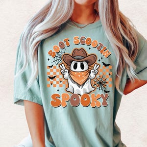 May include: A light blue t-shirt with a graphic of a ghost wearing a cowboy hat and bandana. The ghost is holding up two peace signs. The text "Boot Scootin Spooky" is printed on the shirt.