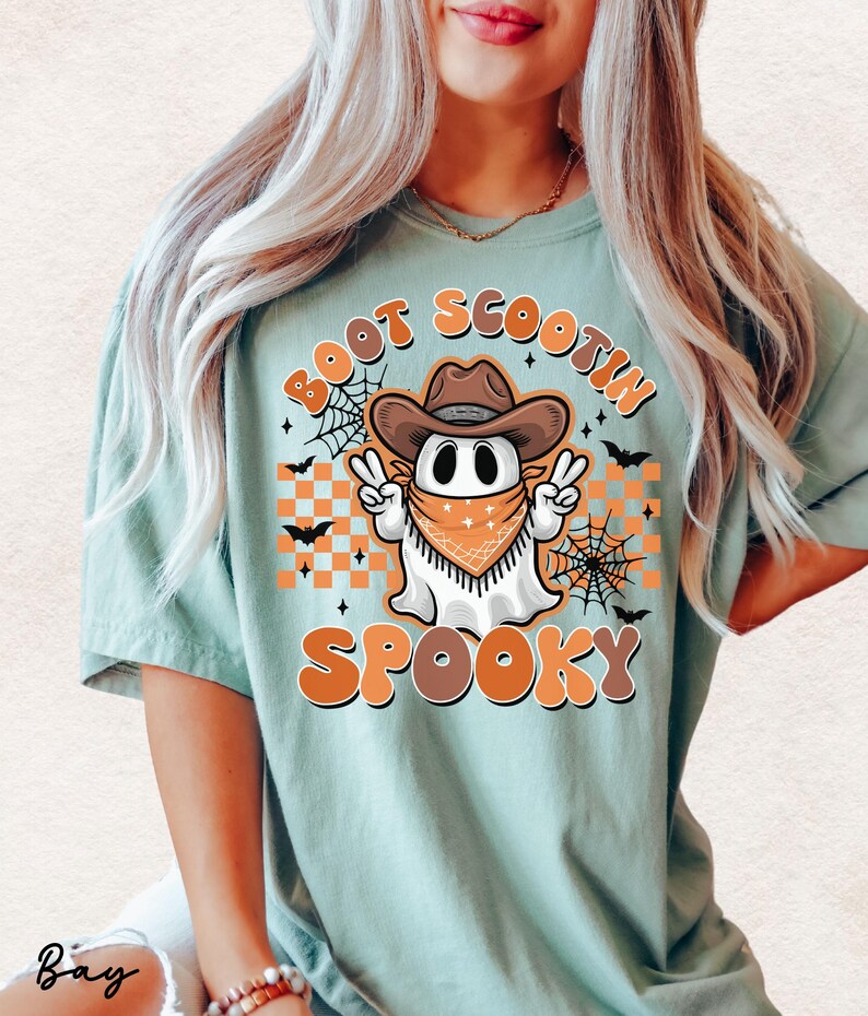 May include: A light blue t-shirt with a graphic of a ghost wearing a cowboy hat and bandana. The ghost is holding up two peace signs. The text "Boot Scootin Spooky" is printed on the shirt.