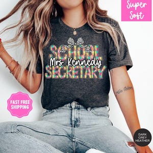 Może przedstawiać: Szary t-shirt w kolorze heather z kolorowym wzorem w kratę, na kt&oacute;rym widnieje napis "School Mrs. Kennedy Secretary" z czarną i białą kokardką w kropki na g&oacute;rze.