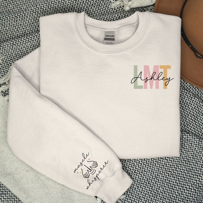 May include: A white sweatshirt with the text "LMT Ashley" on the front and "muscle whisperer" on the left sleeve. The text is in a colourful font.