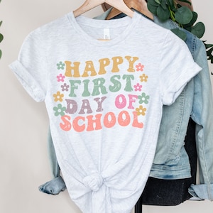 Puede incluir: Una camiseta gris claro con el texto "Happy First Day of School" en letras coloridas y desgastadas, con peque&ntilde;os detalles de flores.