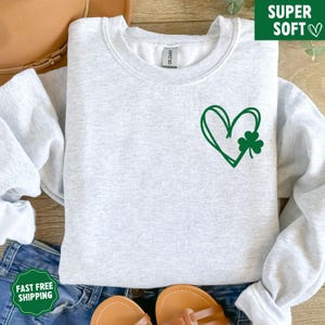 May include: A grey sweatshirt featuring a green heart and shamrock design. The sweatshirt is made from a soft, comfortable material.