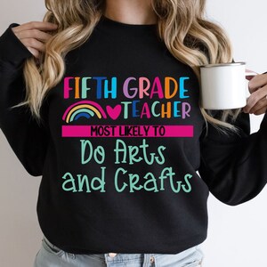 K&ouml;nnte beinhalten: Schwarzer Pullover mit dem Text "Fifth Grade Teacher Most Likely To Do Arts and Crafts" in bunten Buchstaben mit einem Regenbogen und einem Herz-Motiv.