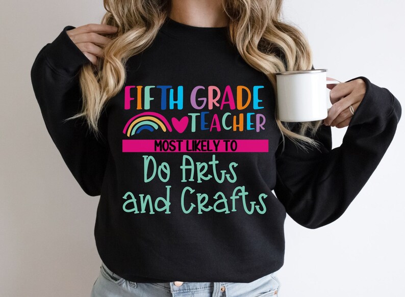 K&ouml;nnte beinhalten: Schwarzer Pullover mit dem Text "Fifth Grade Teacher Most Likely To Do Arts and Crafts" in bunten Buchstaben mit einem Regenbogen und einem Herz-Motiv.