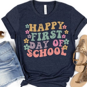 Puede incluir: Una camiseta azul marino con el texto "HAPPY FIRST DAY OF SCHOOL" en una fuente retro con flores coloridas que rodean el texto.