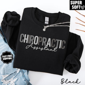 May include: Black crewneck sweatshirt with the words "CHIROPRACTIC Assistant" in gray and white lettering. A silver necklace with a circular pendant is draped on the sweatshirt. The words "SUPER SOFT" are in a black box in the upper right corner.