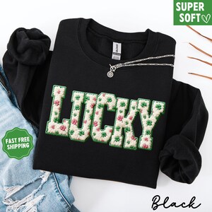 Pu&ograve; includere: Felpa nera con la scritta "LUCKY" ricamata in un carattere strutturato color crema con trifogli verdi e fiori rosa. Una collana d'argento &egrave; drappeggiata sulla felpa. Le parole "SUPER SOFT" sono in una scatola verde.