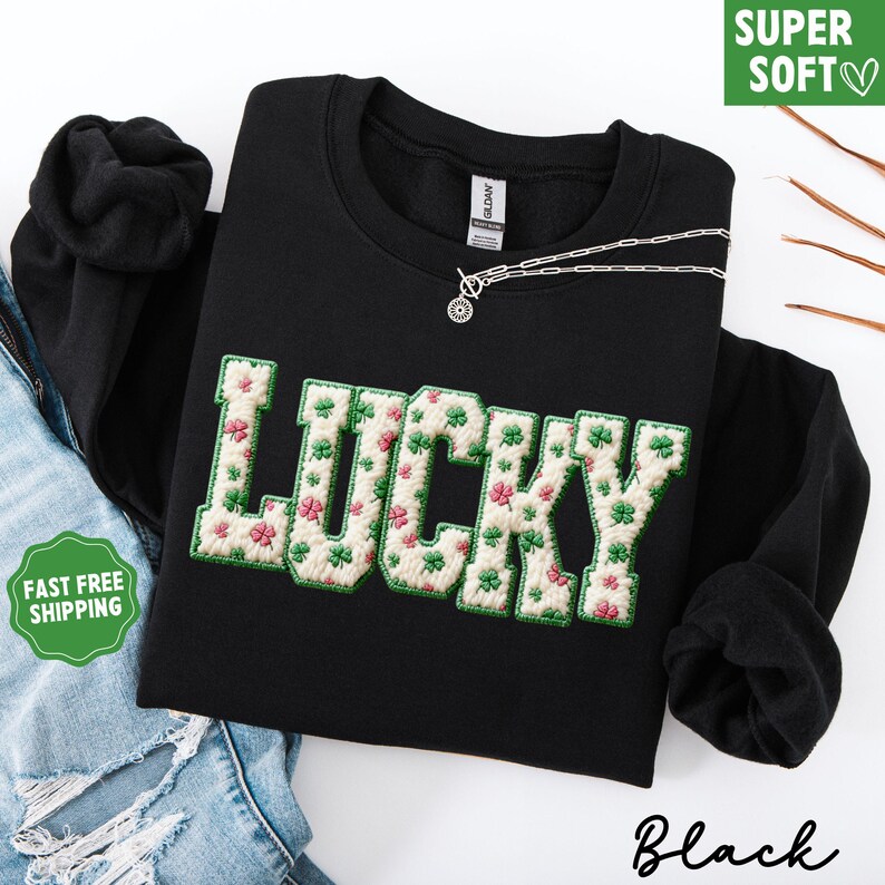 Pu&ograve; includere: Felpa nera con la scritta "LUCKY" ricamata in un carattere strutturato color crema con trifogli verdi e fiori rosa. Una collana d'argento &egrave; drappeggiata sulla felpa. Le parole "SUPER SOFT" sono in una scatola verde.