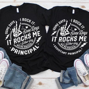 May include: Two black t-shirts with white text and graphics. The text on the shirts reads "Some days I rock it. Some days it rocks me. Either way, we're rockin'" and "Some days I rock it. Some days it rocks me. Either way, we're rockin'" with the words "Principal" and "Assistant Principal" below the text.