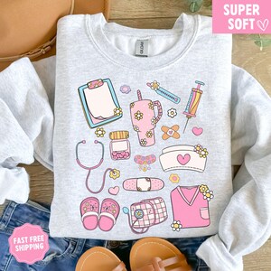 May include: A light gray sweatshirt featuring a collection of pink and pastel medical-themed illustrations. The design includes a stethoscope, syringe, bandages, and a nurse's cap, all adorned with floral accents. The words "SUPER SOFT" are in a pink box.