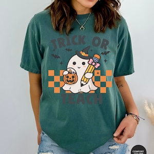 K&ouml;nnte beinhalten: Ein blaugr&uuml;nes T-Shirt mit einem Motiv eines Geistes, der einen Hexenhut tr&auml;gt und einen Bleistift und einen K&uuml;rbis h&auml;lt. Der Text "Trick or Teach" ist auf das Shirt gedruckt.
