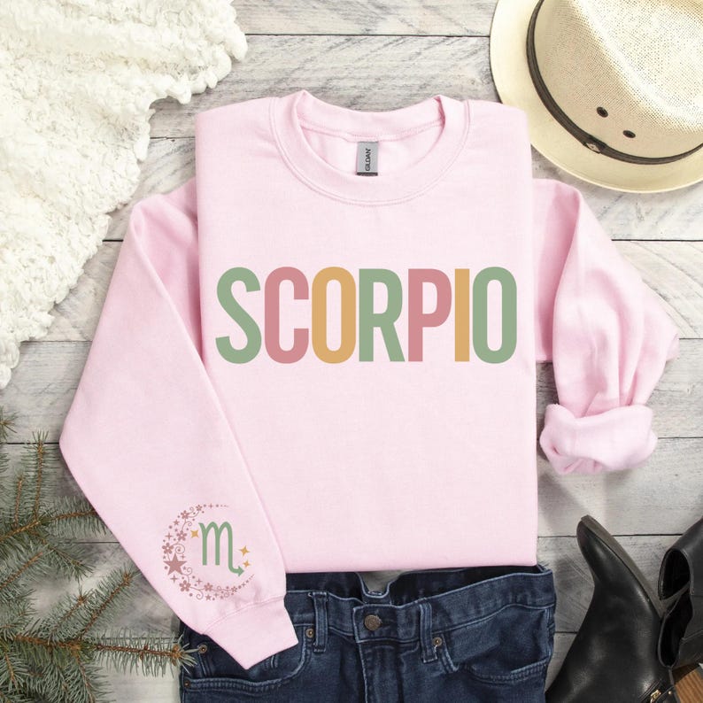 May include: Pink sweatshirt with the word "SCORPIO" in colorful letters. The left sleeve has a small graphic of the Scorpio zodiac symbol.