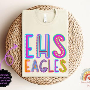 Puede incluir: Una camiseta beige con el texto "EHS Eagles" en letras de colores y contorneadas. Las letras est&aacute;n decoradas con peque&ntilde;os puntos. El texto est&aacute; centrado en la camiseta.