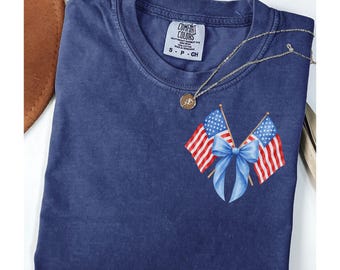 May include: A navy blue t-shirt with a patriotic design. The shirt features two American flags with a blue bow in the center. The text "True Navy" is at the bottom. The shirt has a round neckline and short sleeves.
