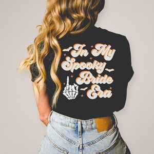 Puede incluir: Camiseta negra con el texto "In My Spooky Bride Era" en naranja y blanco en fuente retro. Una mano de esqueleto blanca con un anillo en el dedo medio est&aacute; por encima del texto.