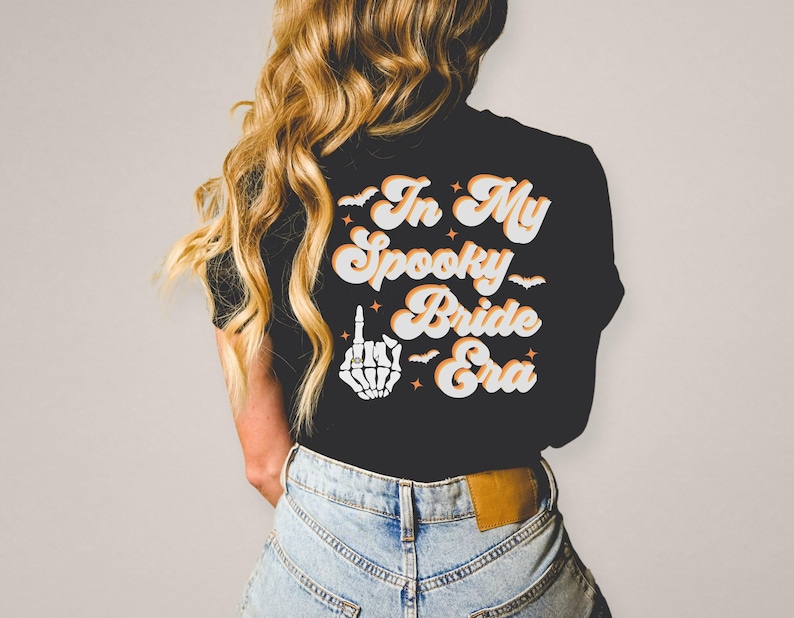 Puede incluir: Camiseta negra con el texto "In My Spooky Bride Era" en naranja y blanco en fuente retro. Una mano de esqueleto blanca con un anillo en el dedo medio est&aacute; por encima del texto.