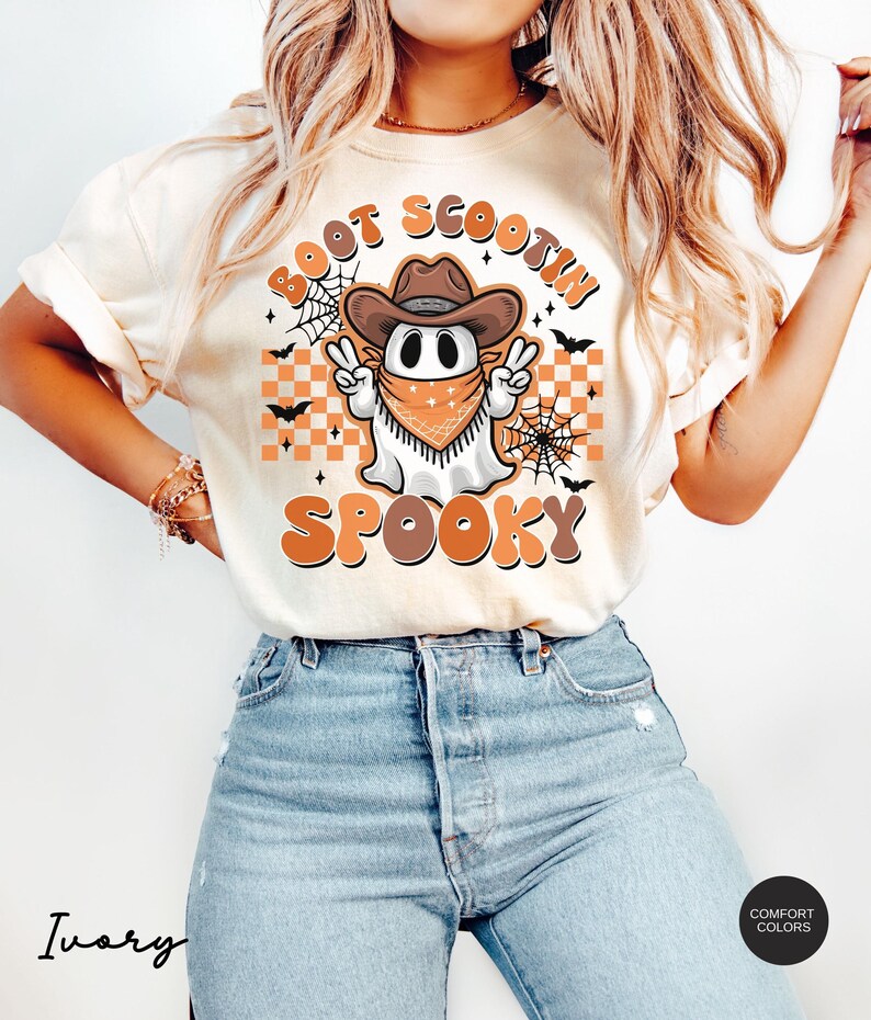 May include: A white t-shirt with an orange and brown graphic of a ghost wearing a cowboy hat and bandana. The text "Boot Scootin Spooky" is written in a retro font around the ghost.