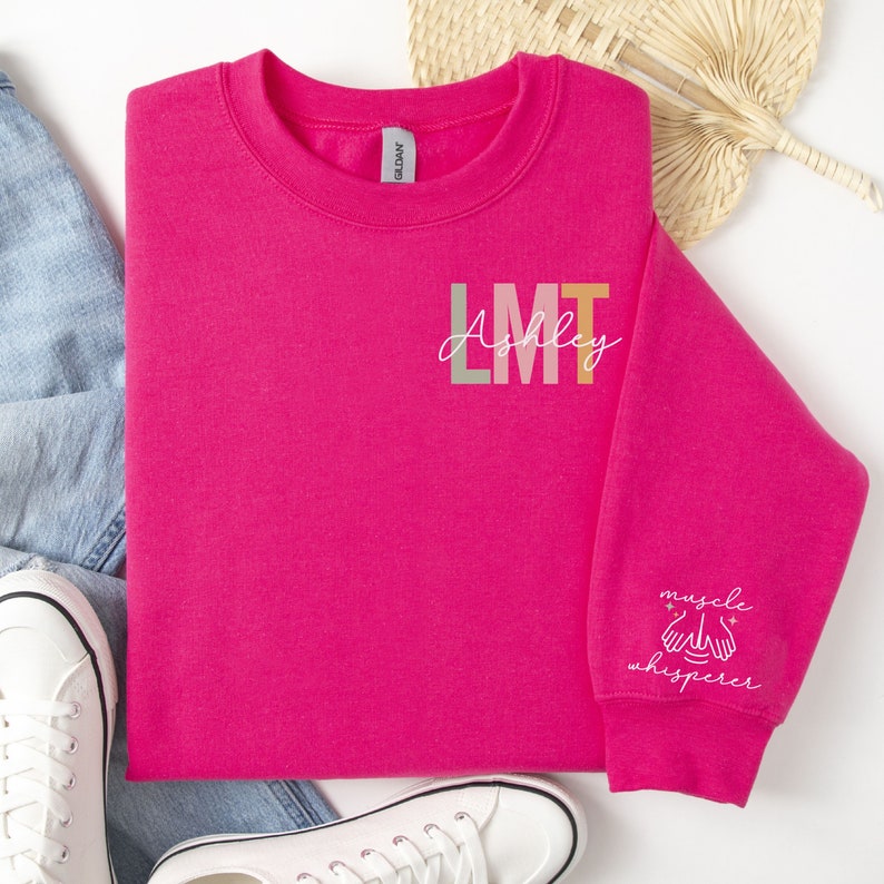 May include: Pink sweatshirt with the text "LMT Ashley" and "muscle whisperer" on the sleeve.