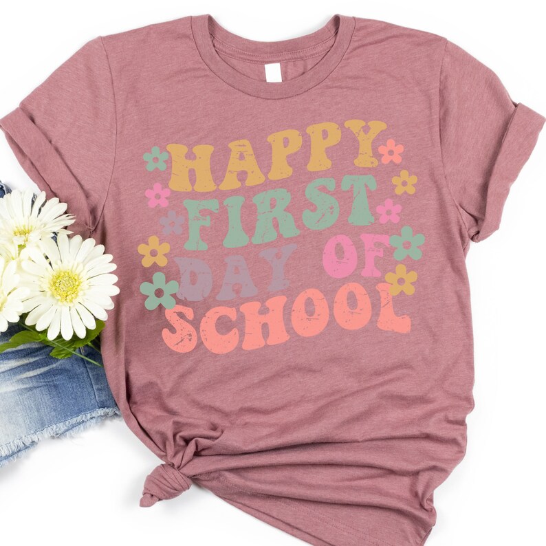 Puede incluir: Una camiseta color malva con el texto "HAPPY FIRST DAY OF SCHOOL" en una fuente retro con flores de colores que rodean el texto.