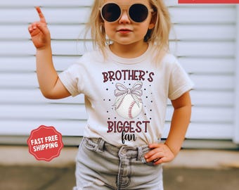 May include: A young girl wearing a white t-shirt with the text "Brother's Biggest Fan" and a baseball graphic. The shirt has a maroon bow and stars. The girl is wearing denim shorts and sunglasses.