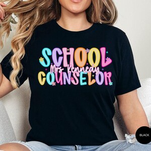 May include: Black t-shirt with the words "SCHOOL Mrs. Kennedy COUNSELOR" in colorful, bubble-style letters. The text is decorated with small flower and star accents. The shirt is a short-sleeved crew neck.