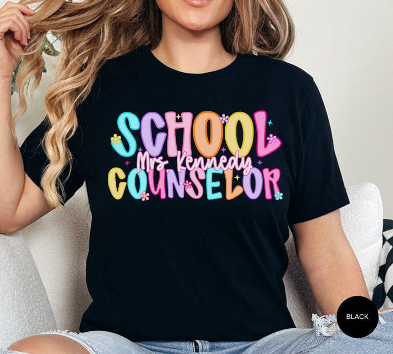 May include: Black t-shirt with the words "SCHOOL Mrs. Kennedy COUNSELOR" in colorful, bubble-style letters. The text is decorated with small flower and star accents. The shirt is a short-sleeved crew neck.