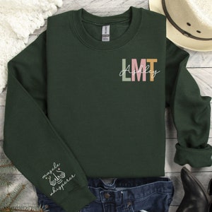 May include: A dark green sweatshirt with the text "LMT Ashley" in colourful letters on the front. The left sleeve has the text "muscle whisperer" with a star and a hand symbol.