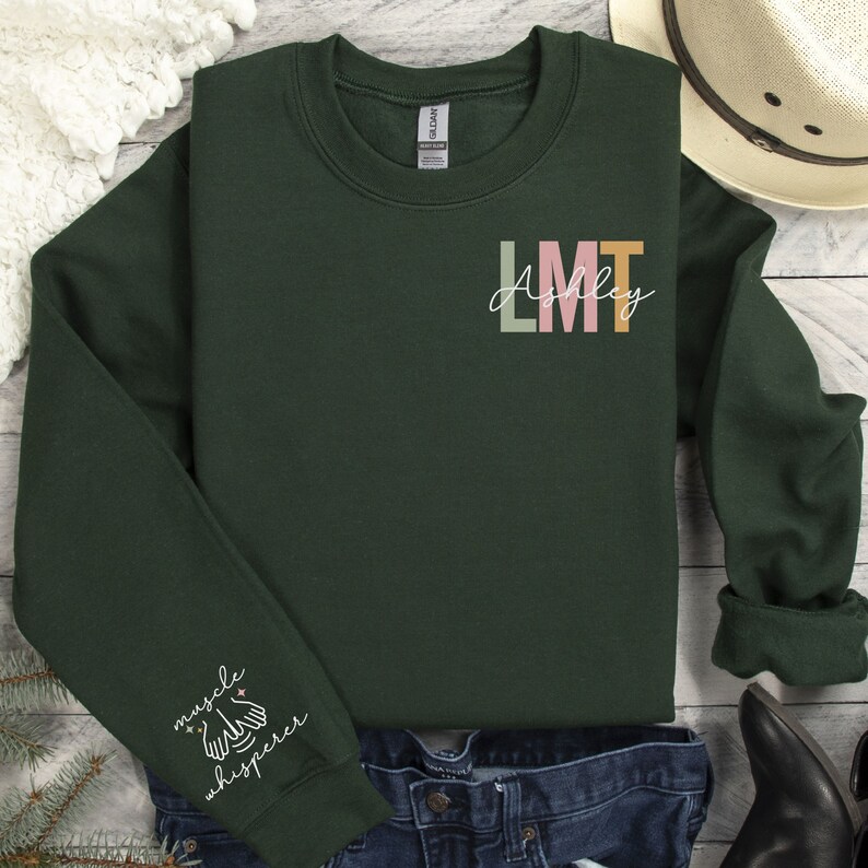 May include: A dark green sweatshirt with the text "LMT Ashley" in colourful letters on the front. The left sleeve has the text "muscle whisperer" with a star and a hand symbol.