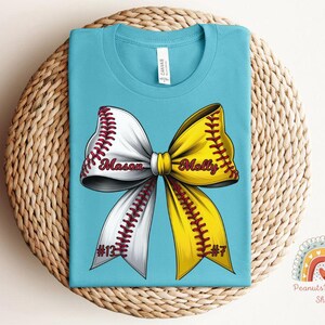 May include: A turquoise blue t-shirt with a white and yellow softball bow. The bow has red stitching and the names "Mason #13" and "Molly #7" are written on the bow.