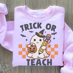 May include: Pink sweatshirt with a ghost holding a pencil and a pumpkin. The text "Trick or Teach" is printed on the sweatshirt.