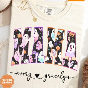 May include: Ivory-colored t-shirt with the word "MAMA" in large, colorful letters filled with ghost and pumpkin graphics. The names "avery" and "gracelyn" are written below the word "MAMA". A gold necklace is draped on the shirt.