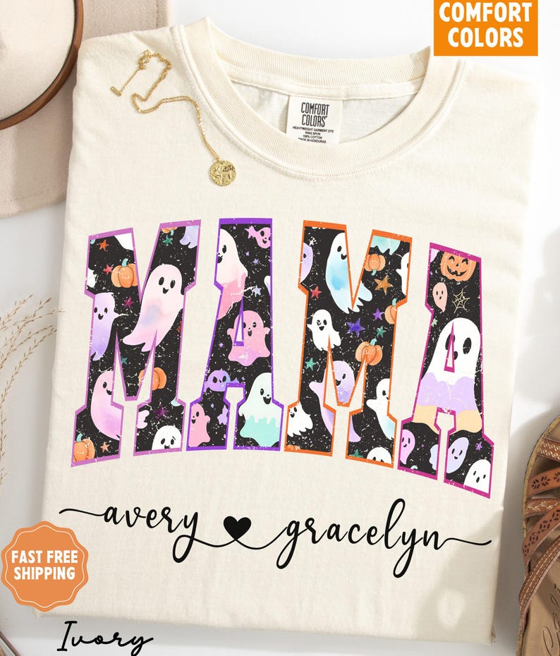 May include: Ivory-colored t-shirt with the word "MAMA" in large, colorful letters filled with ghost and pumpkin graphics. The names "avery" and "gracelyn" are written below the word "MAMA". A gold necklace is draped on the shirt.