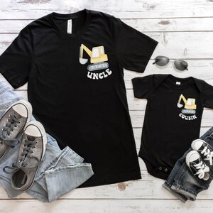 Peut inclure: T-shirt noir avec un dessin de pelle m&eacute;canique jaune et le mot "UNCLE". Un body de b&eacute;b&eacute; noir assorti avec un motif de pelle m&eacute;canique et le mot "COUSIN". Baskets grises, jean bleu et lunettes de soleil.