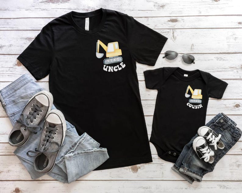 Peut inclure: T-shirt noir avec un dessin de pelle m&eacute;canique jaune et le mot "UNCLE". Un body de b&eacute;b&eacute; noir assorti avec un motif de pelle m&eacute;canique et le mot "COUSIN". Baskets grises, jean bleu et lunettes de soleil.