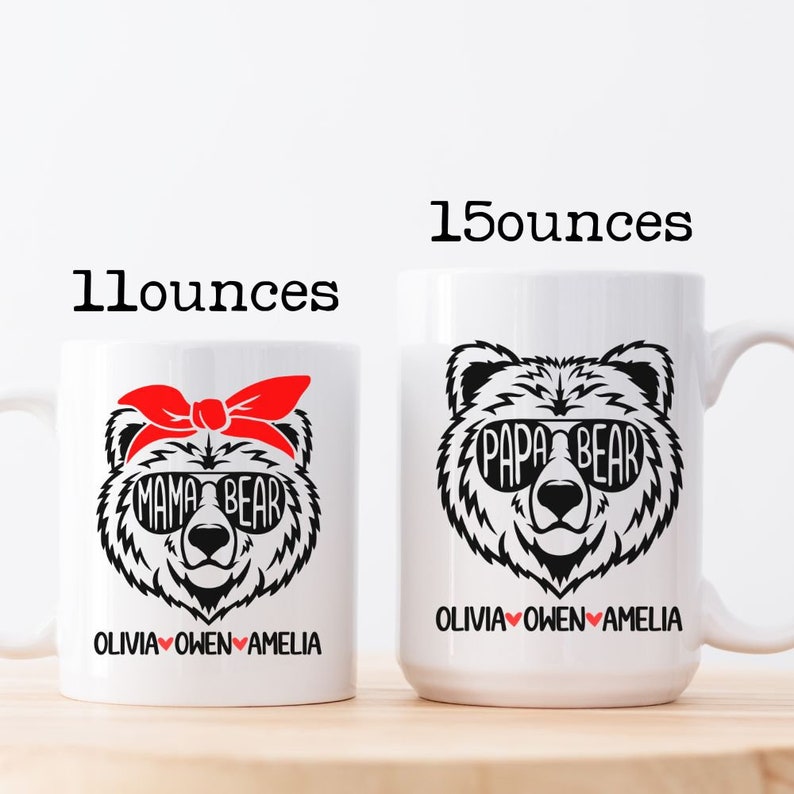 Op de afbeelding: Twee witte keramische mokken met zwarte beerillustraties. De kleinere mok is 11 ounce en heeft een mamabeer met een rode bandana en zonnebril. De grotere mok is 15 ounce en heeft een papabeer met zonnebril. Beide mokken hebben de tekst "Mama Bear" en "Papa Bear" en de namen "Olivia, Owen, Amelia" gedrukt op de bodem.