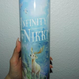 Puede incluir: Un vaso de acero inoxidable azul y blanco con un gráfico de un ciervo blanco en un entorno de bosque. El vaso tiene el texto "INFINITY NIKKI" impreso en él.
