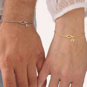 May include: Two matching bracelets, one silver and one gold, each with a small initial charm. The silver bracelet has the letter "A", and the gold bracelet has the letter "H". Both bracelets are on wrists, with the hands clasped together.