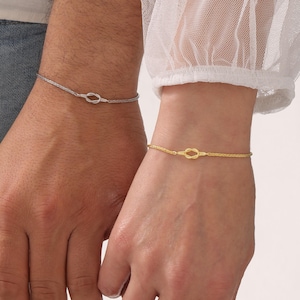 Puede incluir: Dos pulseras a juego, una plateada y otra dorada, cada una con un diseño de nudo. Las pulseras se llevan en las muñecas, mostrando un estilo sencillo y elegante. La pulsera dorada está en la muñeca derecha, y la plateada en la izquierda.