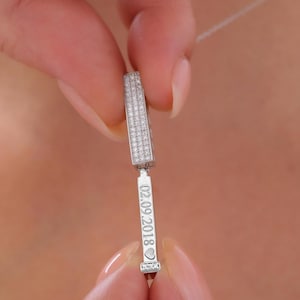 Op de afbeelding: Een zilveren hangerketting met een rechthoekig bovendeel bedekt met kleine, heldere stenen. Het onderste gedeelte heeft de datum "02.09.2018" gegraveerd, met een hartsymbool aan het einde. De ketting wordt tussen twee vingers gehouden.