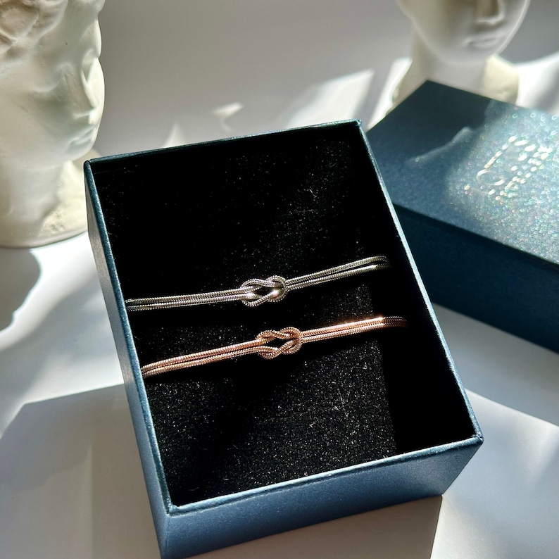 May include: Two knot bracelets, one silver and one rose gold, displayed in a blue box.