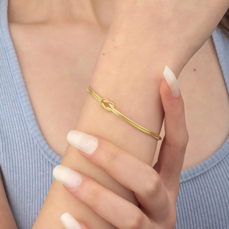 May include: A gold knot bracelet with a delicate design. The bracelet is made of two thin strands of gold that are twisted together to form a knot.