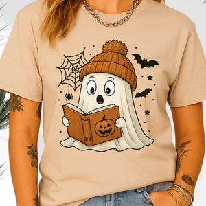 May include: Beige t-shirt featuring a cartoon ghost wearing a brown beanie and reading a book with a jack-o'-lantern on the cover. The design includes a spider web, bats, and stars, perfect for Halloween.