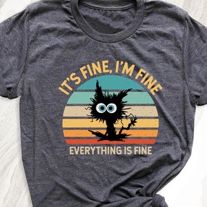 May include: A dark gray t-shirt with a vintage-style graphic of a black cat with big blue eyes. The graphic says "It's Fine, I'm Fine, Everything is Fine".