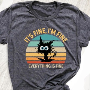 May include: A dark grey t-shirt with a vintage-style sunset graphic featuring a black cat with large blue eyes. The text "It's fine, I'm fine, everything is fine" is printed on the shirt.