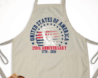May include: A cream-coloured apron with two front pockets and a neck strap. The apron has a circular graphic with the text "UNITED STATES OF AMERICA" and "250th ANNIVERSARY 1776-2026" with an image of the Statue of Liberty. A loaf of bread rests on a burlap cloth.