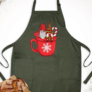 May include: Dark green apron with two front pockets and black ties. A red mug graphic with a snowflake and three gnomes wearing hats and holding a candy cane is centered on the apron. A loaf of bread sits on a burlap cloth.