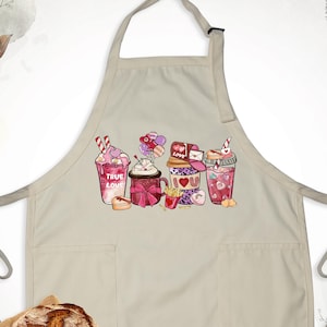 May include: Beige apron with a colorful graphic of Valentine's Day themed drinks, including a pink milkshake with the text "TRUE LOVE". The apron has two pockets and a bread loaf on a burlap cloth.
