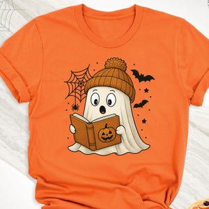 May include: Orange t-shirt featuring a cartoon ghost wearing a brown beanie and reading a book with a pumpkin on the cover. The design includes bats, stars, and a spider web, perfect for Halloween.
