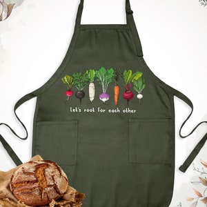 May include: Green kitchen apron with a row of colorful root vegetables and the text "Let's root for each other".
