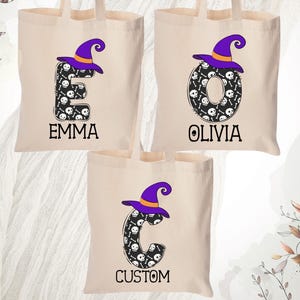 May include: Beige canvas tote bags with a Halloween theme. Each bag features a letter with a witch hat, skull and crossbones design. The names "EMMA", "OLIVIA", and "CUSTOM" are printed below the letters. The bags have long handles.
